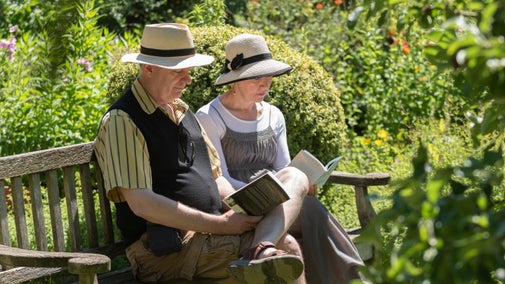 Two visitors sit on a bench in the garden at Wordsworth House to read their books. It's a sunny day in early summer and the couple are surrounded by green foliage